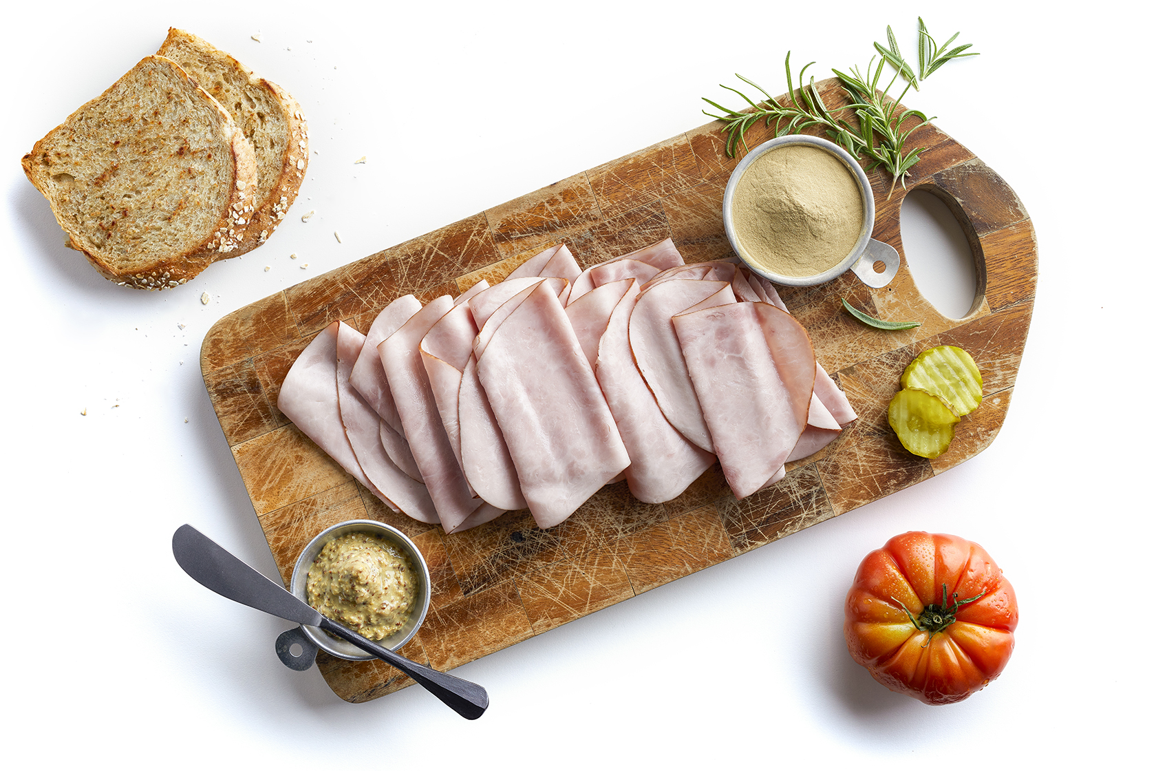 Sliced ham on a cutting board with bread, mustard, and tomato. 