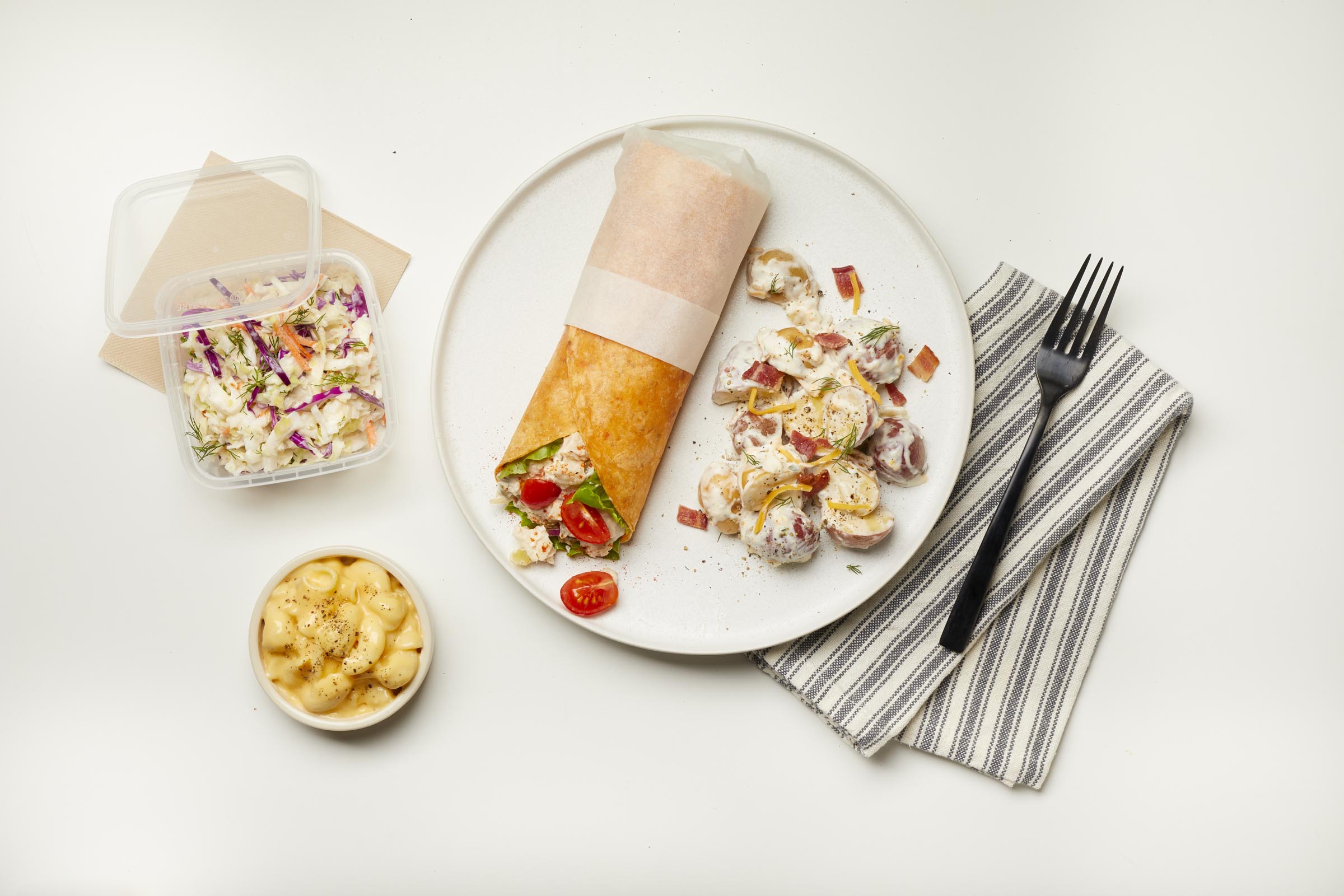 Plate with a wrap, potato salad, coleslaw in a container, and mac and cheese in a bowl on a table.
