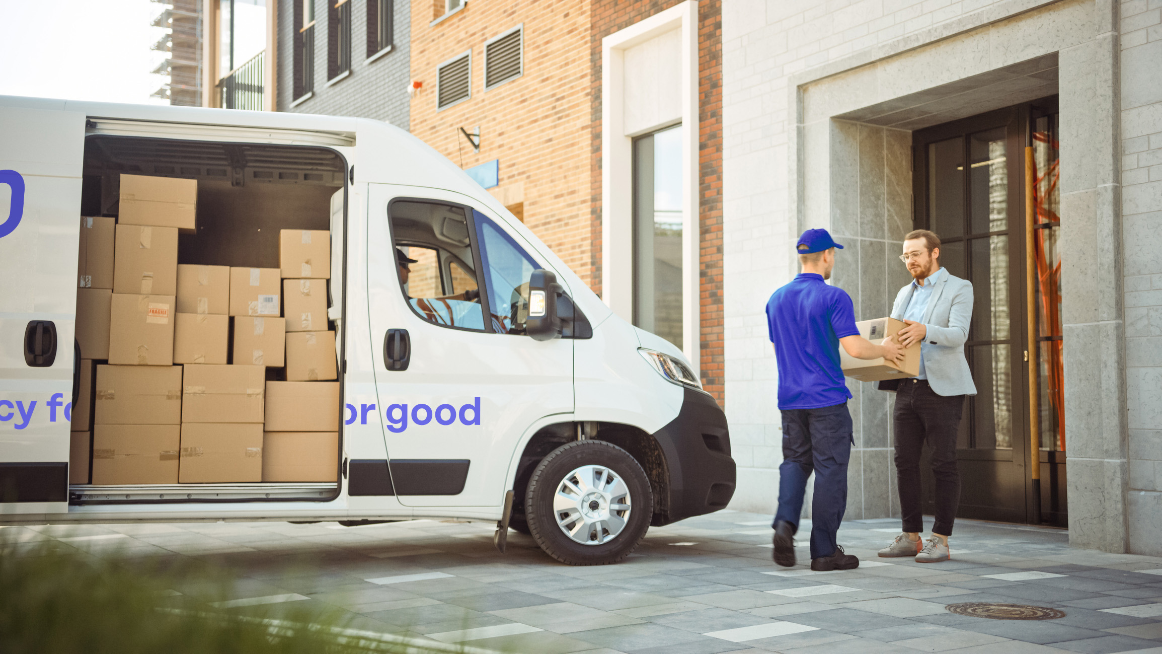 There is a mail truck full of parcels parks at the doorstep. The postman in blue hands over a parcel to the man in the grey suit.