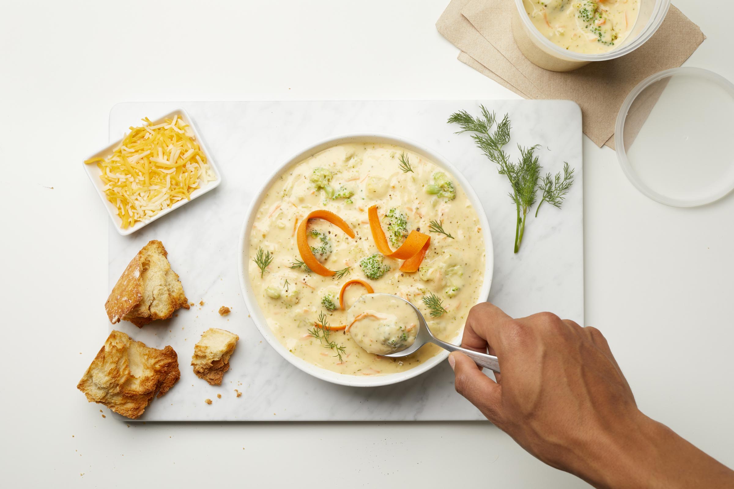 Bowl of creamy broccoli cheddar soup with dill, carrot ribbons, and bread on a marble surface.